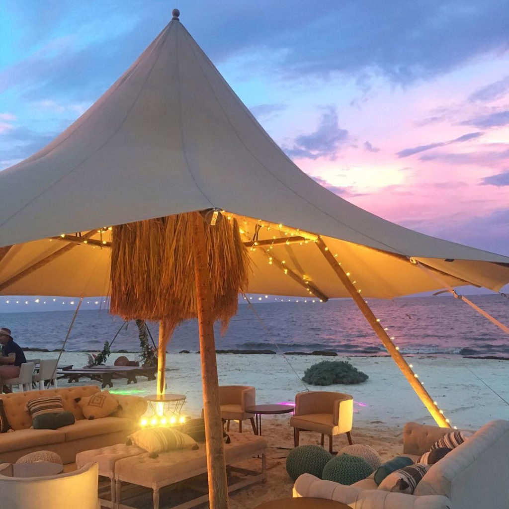 Illuminated Tipi on a sandy beach at sunset, with fairy lights and seating, and the sea and a colourful sky behind it
