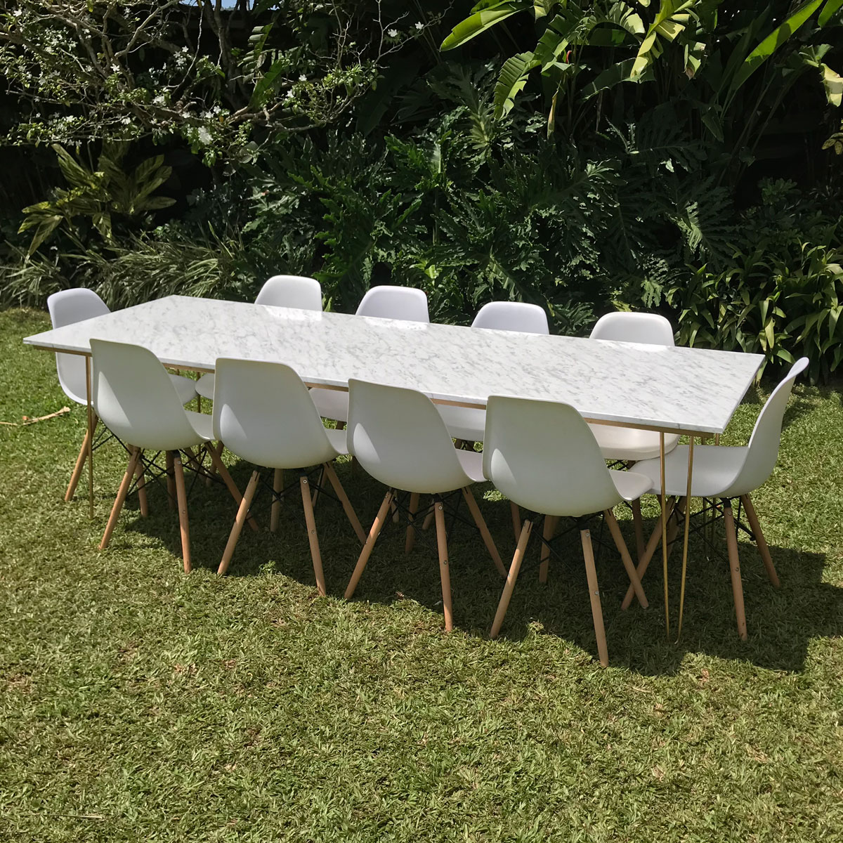 A Marble Hairpin Table with ten white chairs from Trinidad Event Furniture is arranged on green grass, surrounded by lush plants. - Perched Event Rentals, Trinidad