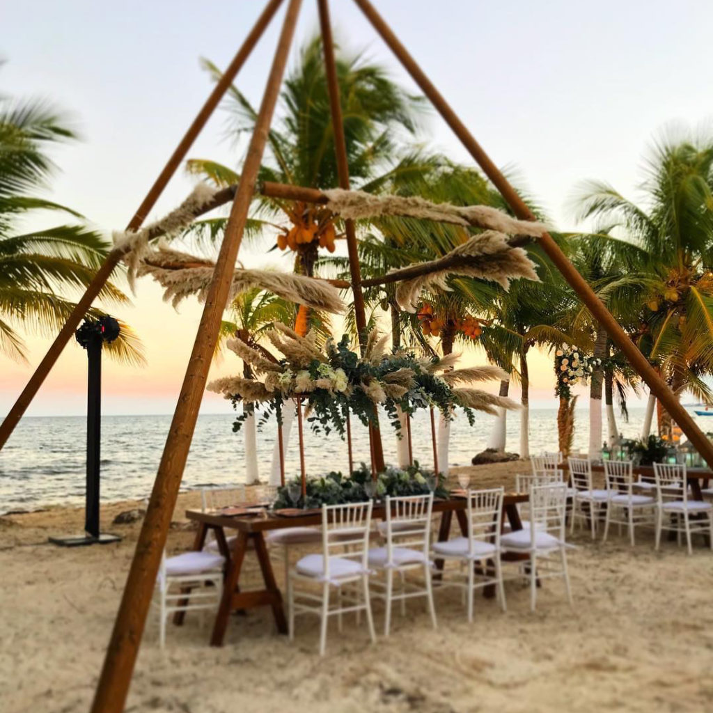 Naked Tipi with a bamboo canopy above wooden tables and white chairs on a beach near palm trees and the sea at sunset