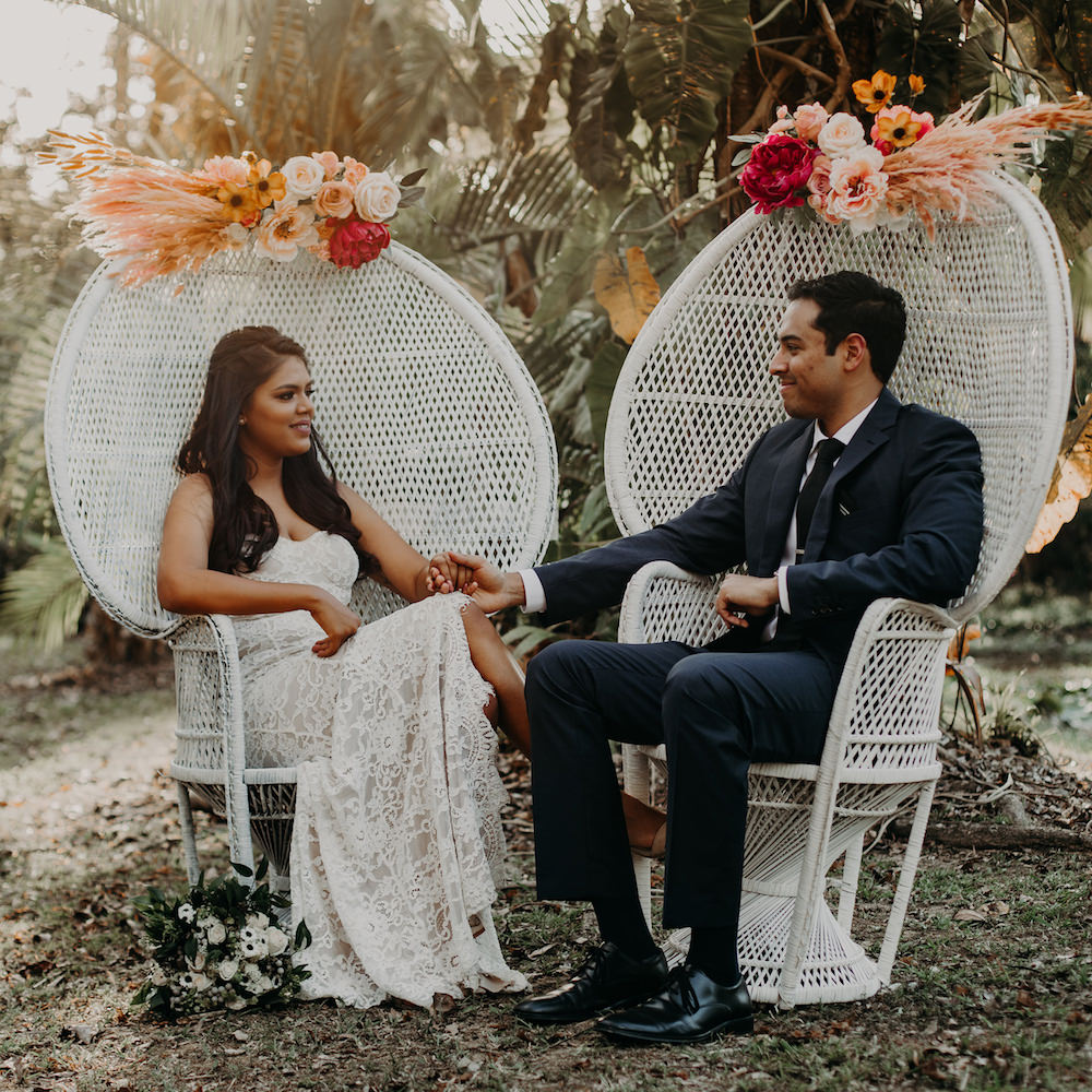 A bride and groom sit outdoors in Peacock Chairs from Trinidad Wedding Hire, holding hands and facing one another, with floral arrangements adorning the chairs. - Perched Event Rentals, Trinidad