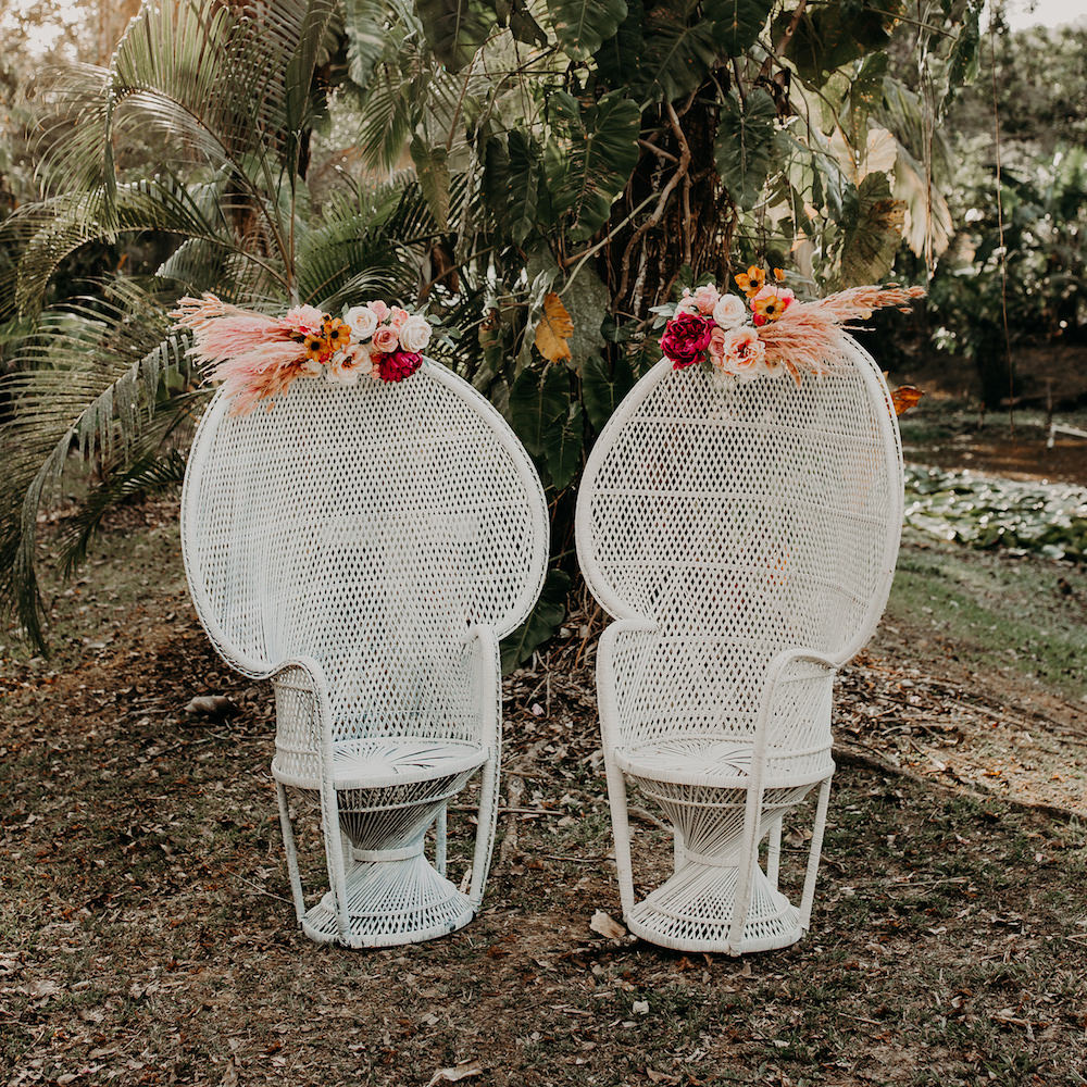Two white Peacock Chairs from Trinidad Event Furniture, featuring tall rounded backs, are adorned with colourful floral arrangements at the top and set outdoors among greenery and trees. - Perched Event Rentals, Trinidad