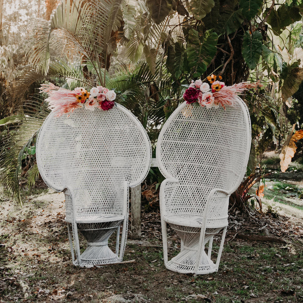Two white Peacock Chairs with pink and red floral arrangements sit side by side outdoors among tropical plants—an elegant seating option from Trinidad Wedding Hire. - Perched Event Rentals, Trinidad