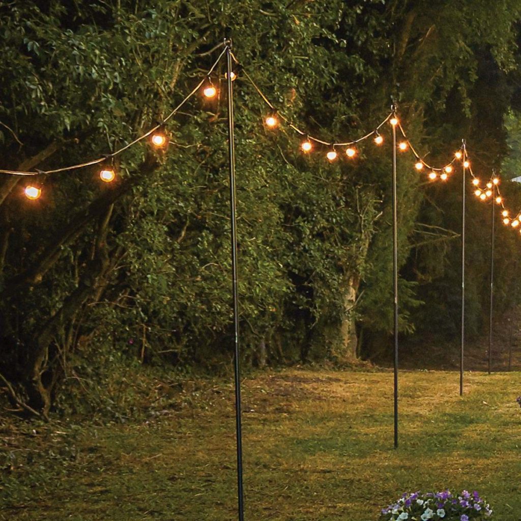 String Light Poles with string lights hanging between them over a grassy area bordered by dense trees, flowers in the corner