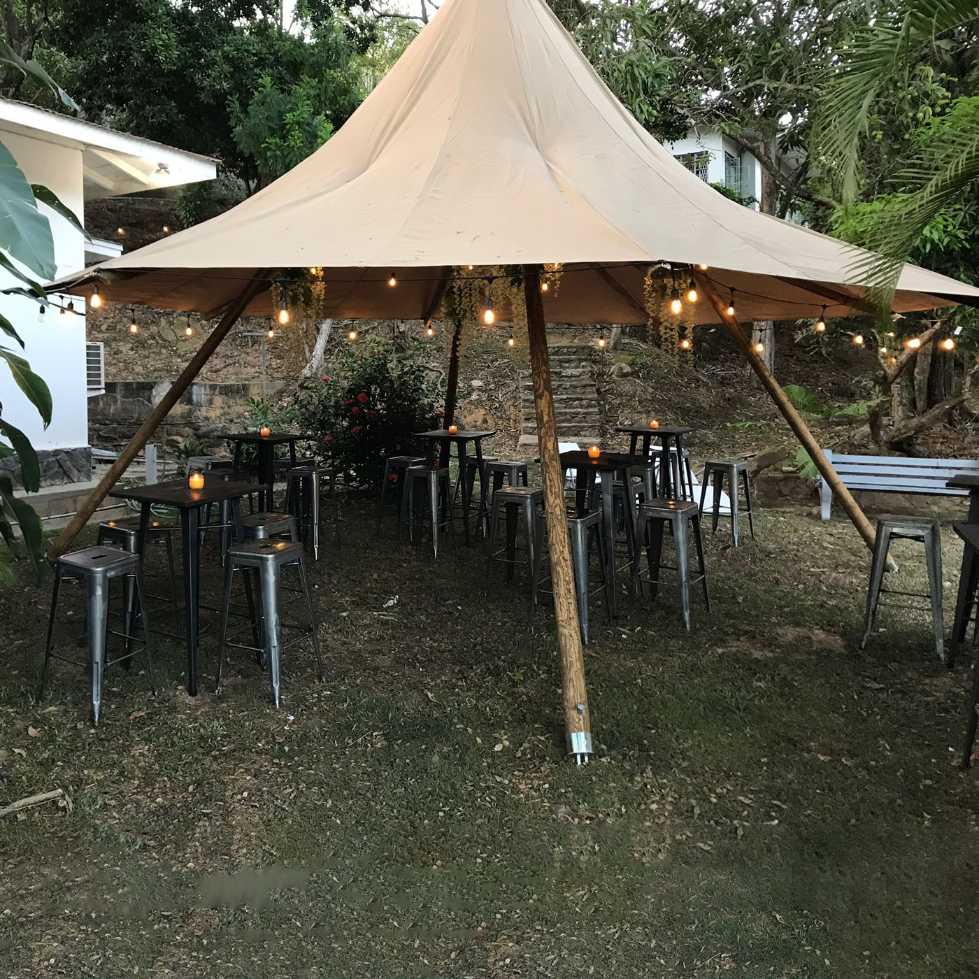 A beige teepee tent covers metal tables and stools from Trinidad Event Hire, with hanging fairy lights, set up outdoors on grass near trees and buildings. - Perched Event Rentals, Trinidad