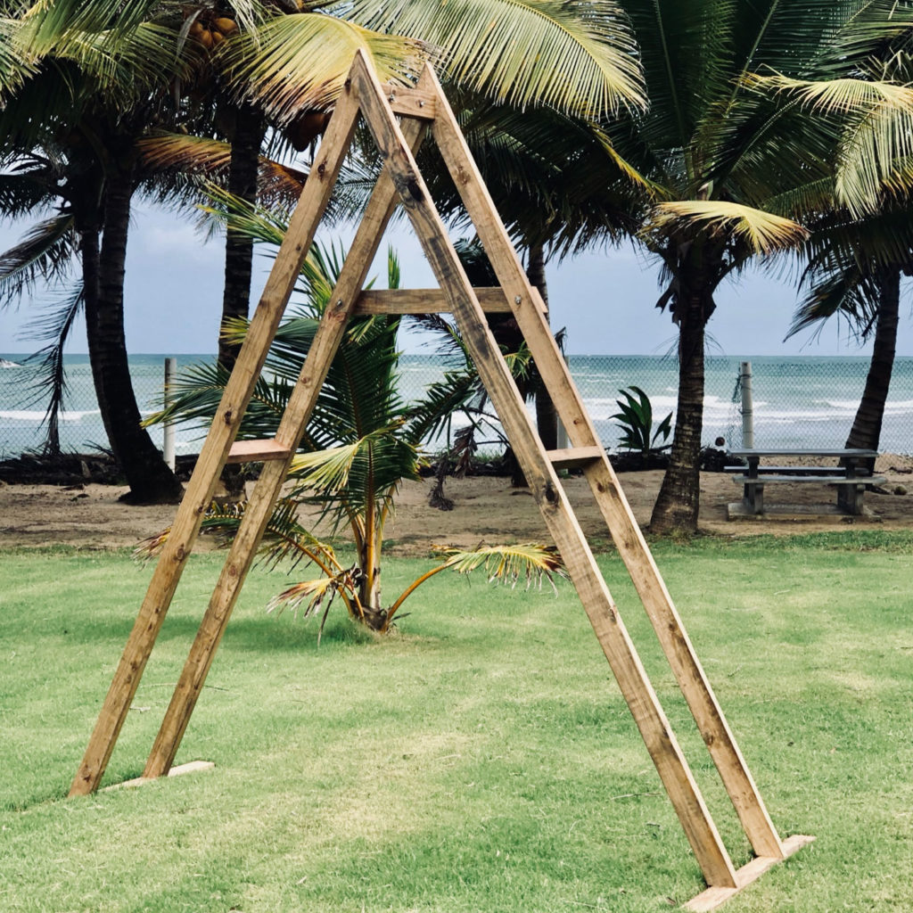 An A-Frame Arch stands on grass with palm trees and the sea behind it