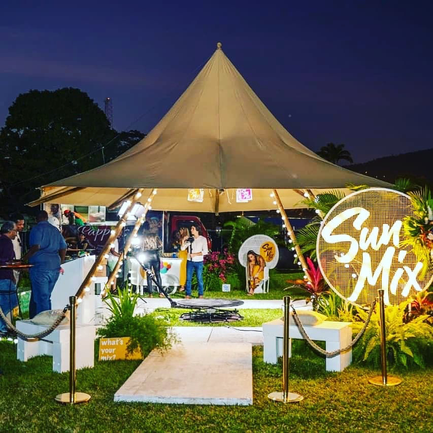 Tipi Tent with a pointed roof, lit at night for an event, with decorative plants and people gathered around it