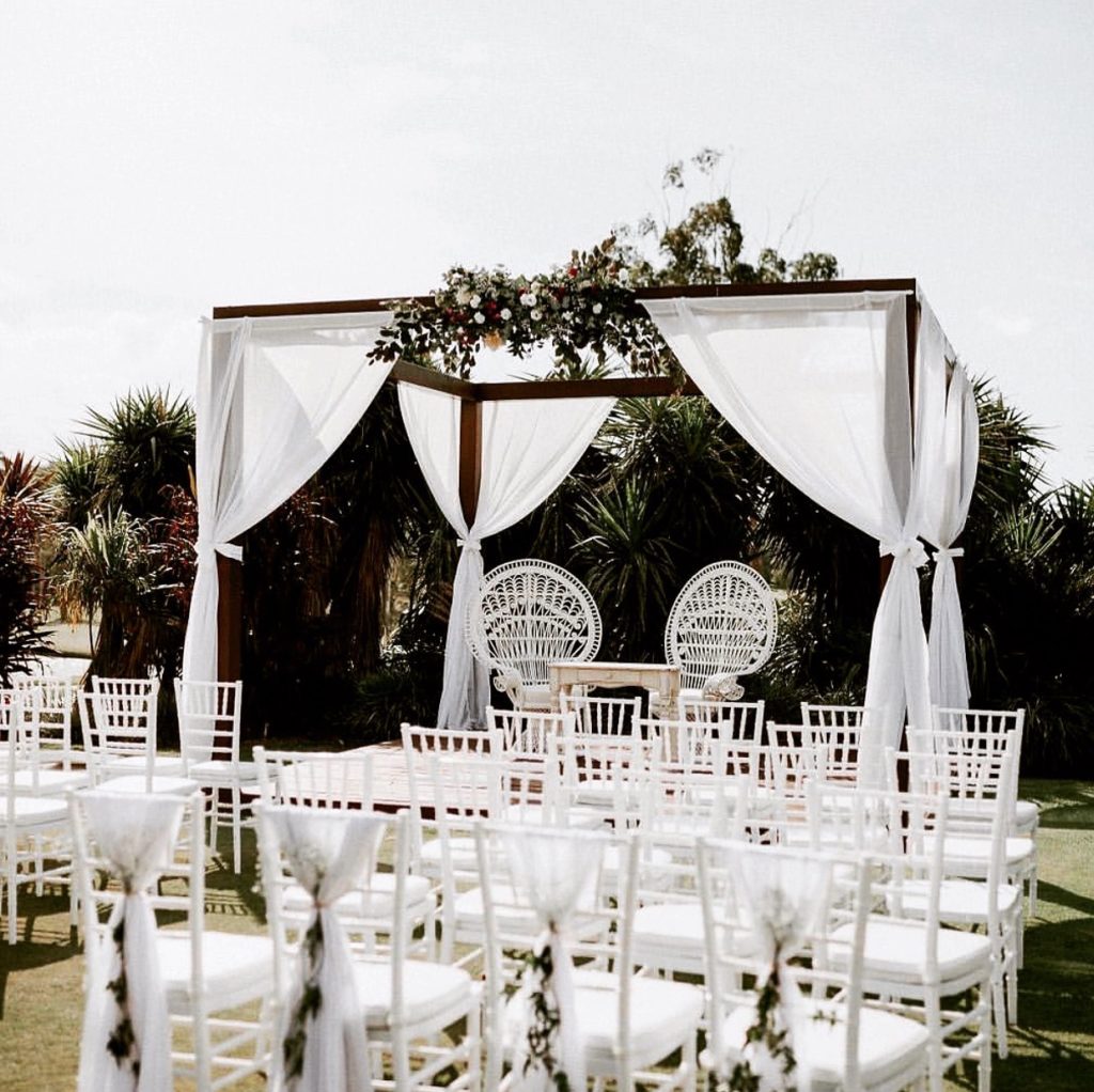 The Pergola Arch with drapes at an outdoor wedding set-up, with white chairs, two decorative chairs beneath it and greenery