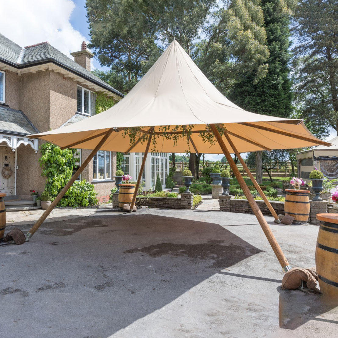 A beige canopy tent with wooden supports, available from Trinidad Event Hire, is set up on a paved area in front of a house surrounded by greenery and decorative barrels. - Perched Event Rentals, Trinidad
