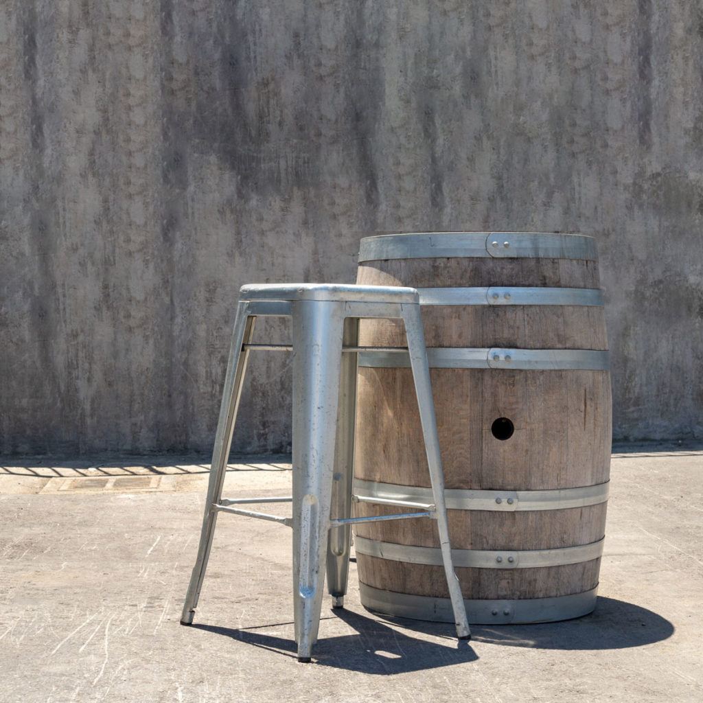 Rum Barrel, a wooden barrel with metal bands, beside a metal stool on concrete against a grey textured wall