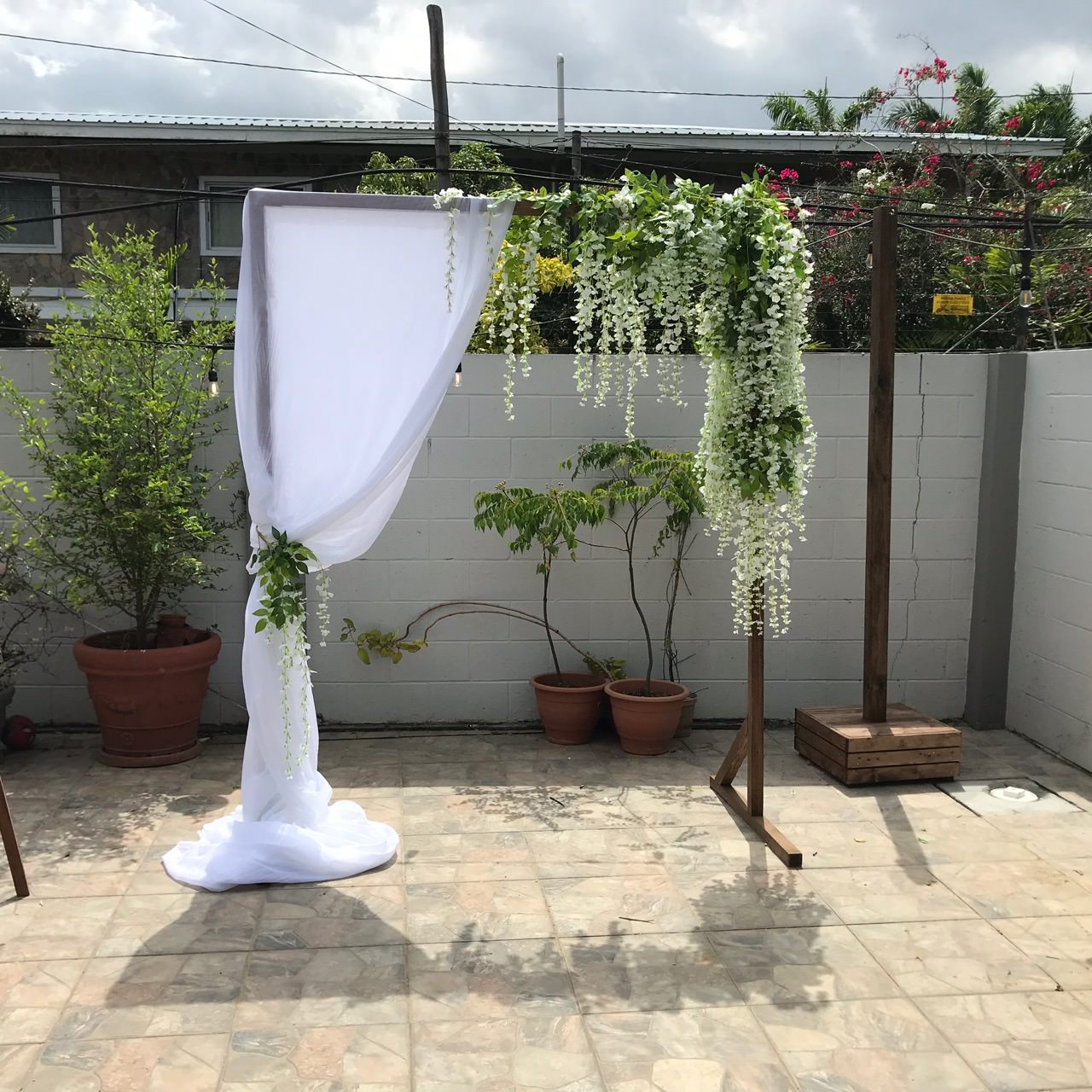 A wooden wedding arch with white drapery and hanging white flowers from Trinidad Wedding Hire stands on a tiled patio beside potted plants and a white brick wall. - Perched Event Rentals, Trinidad