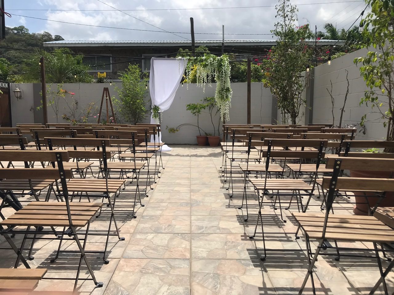 Rows of wooden chairs from Trinidad Event Furniture are arranged outdoors, facing a small wedding altar with white fabric and greenery on a tiled patio, surrounded by walls and plants. - Perched Event Rentals, Trinidad