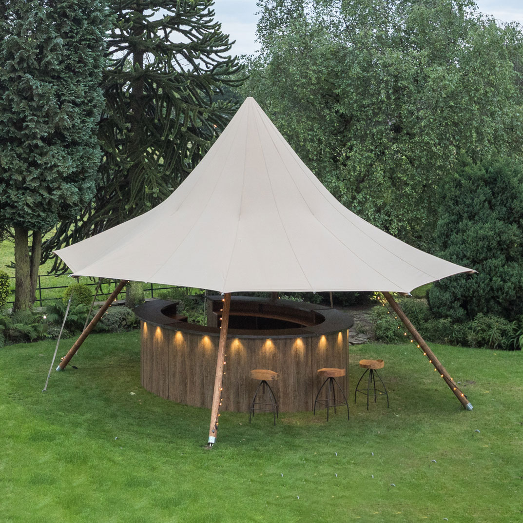 A circular outdoor bar with a beige canopy roof and wooden supports, surrounded by green grass and trees, with two bar stools at the counter—perfect for any occasion from Trinidad Event Hire. - Perched Event Rentals, Trinidad