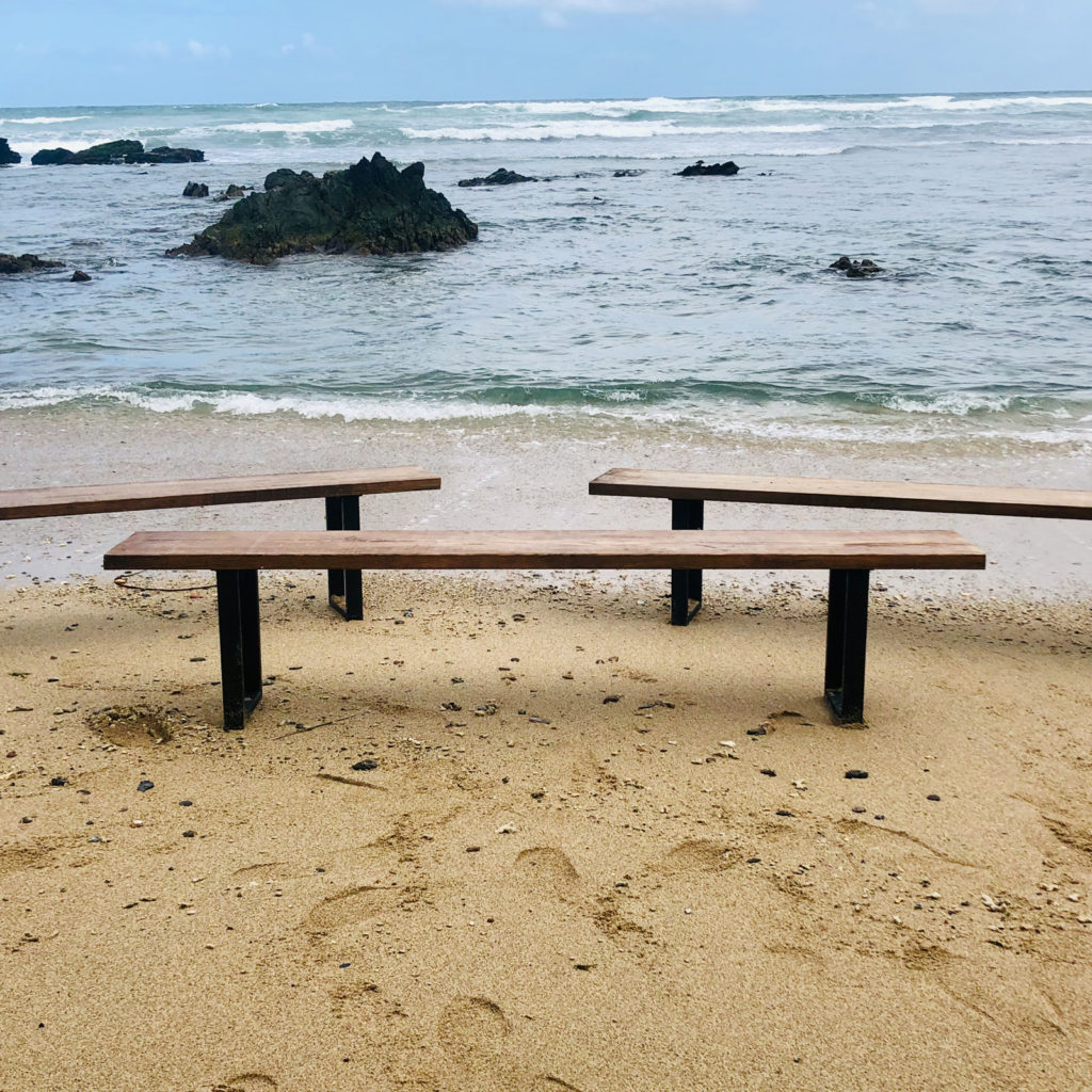 Three French Farm Benches with metal legs set on a sandy beach facing the sea, with rocks and waves in the background